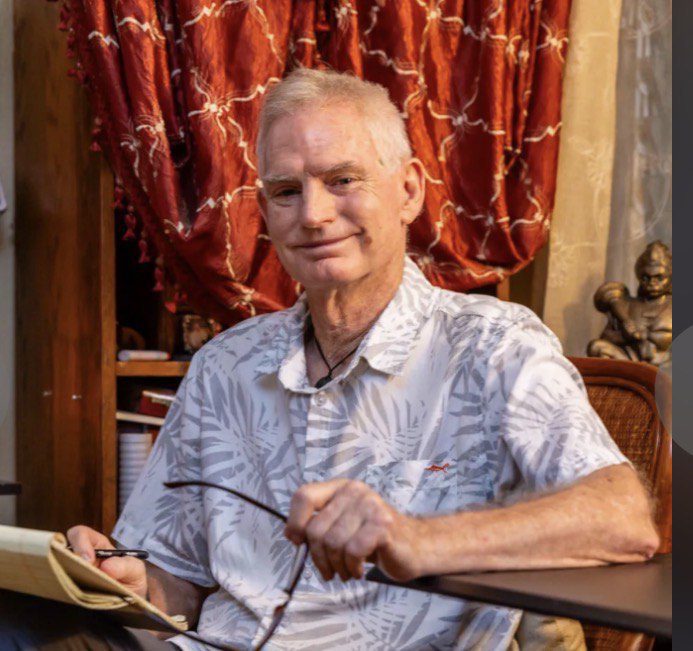 An older man wearing glasses and a patterned shirt sits with a book and pen.